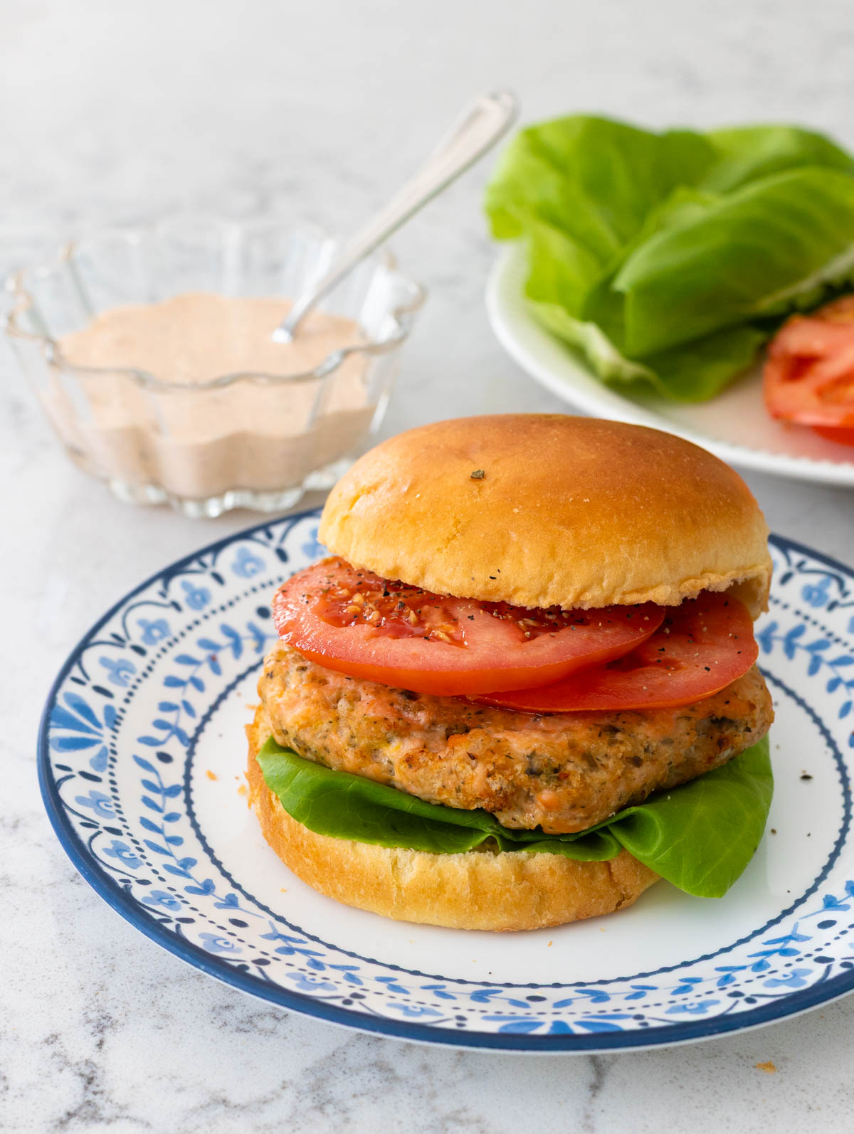 The plain burger is on a plate, a cup of tartar sauce with a spoon is in the background.