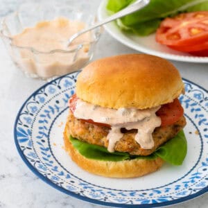The salmon burger is on a brioche bun with lettuce and tomato. A drizzle of tartar sauce drips down the side.