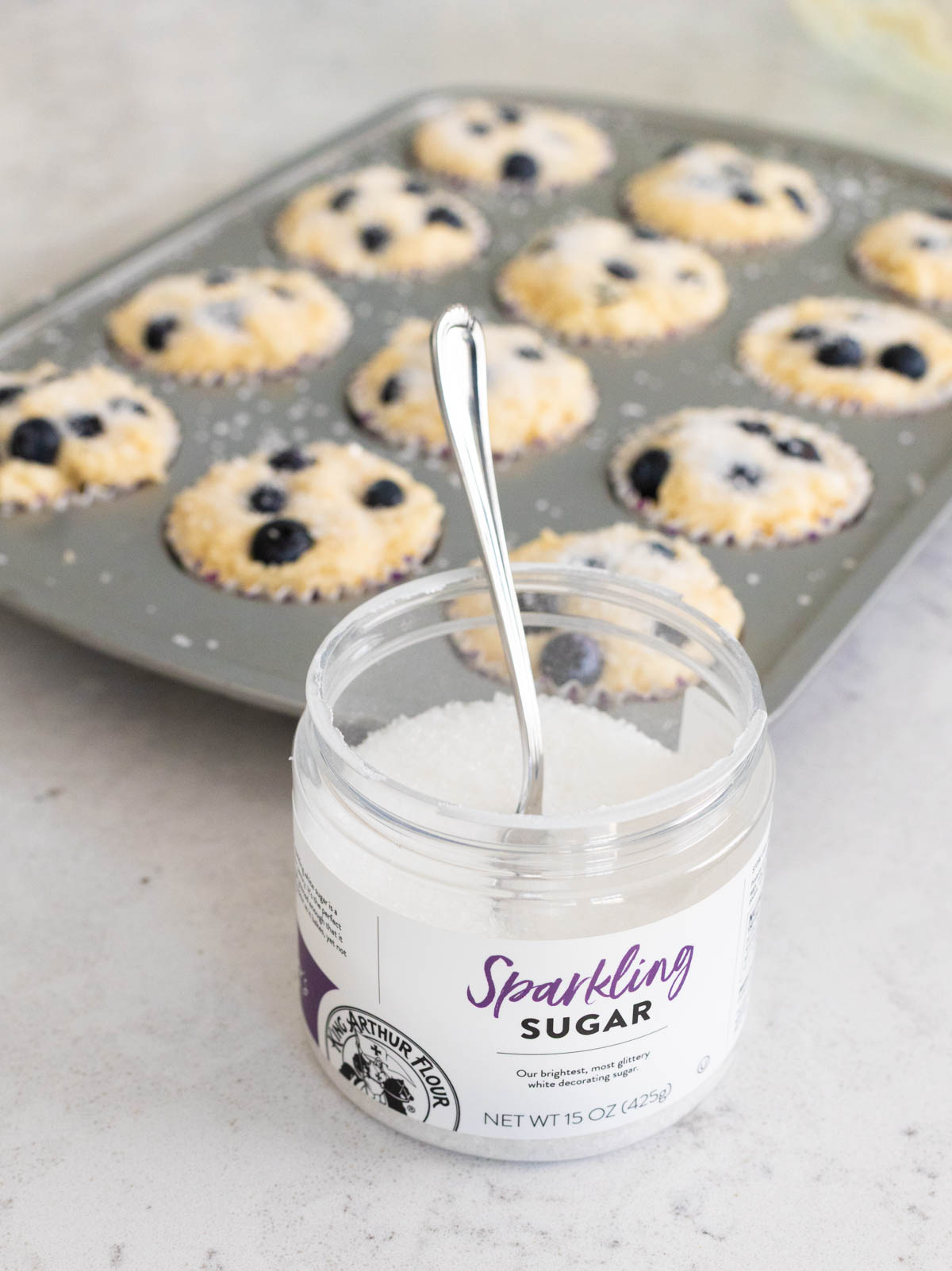 A container of sparkling sugar sits in front of a muffin tin filled with blueberry muffin battter.