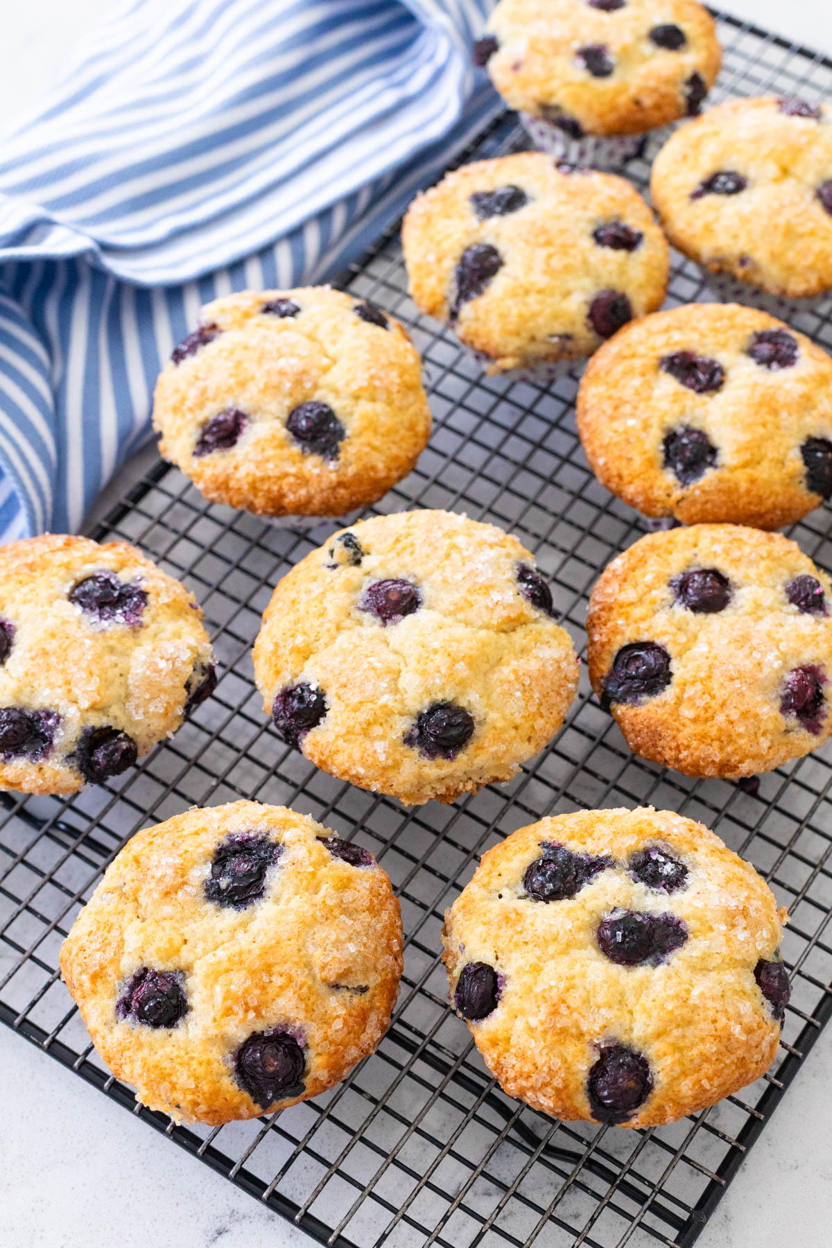 A batch of blueberry muffins with a sparkly sugar topping are cooling on a baker's rack next to a blue and white striped towel.