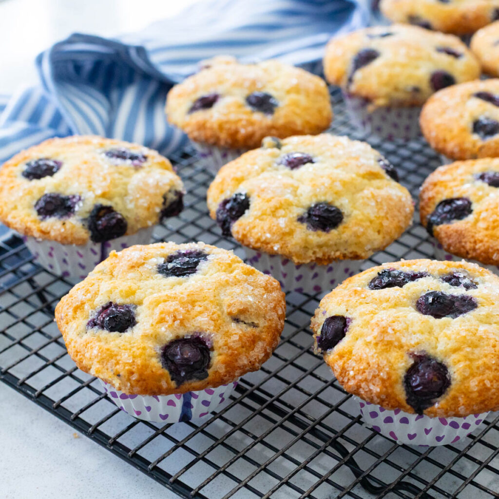 A batch of blueberry muffins with a sparkly sugar topping are cooling on a baker's rack next to a blue and white striped towel.