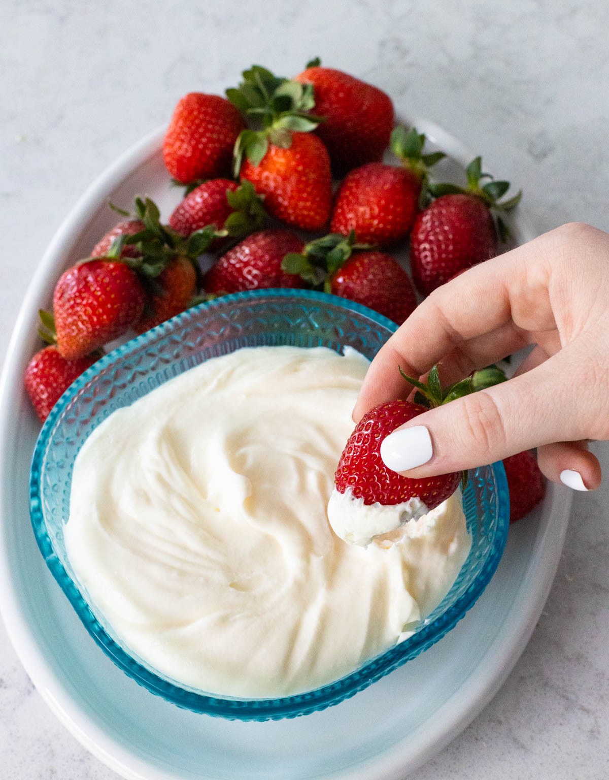 A blue bowl is filled with a 2-ingredient fruit dip on a platter of fresh strawberries. A hand is dipping a berry into the dip.