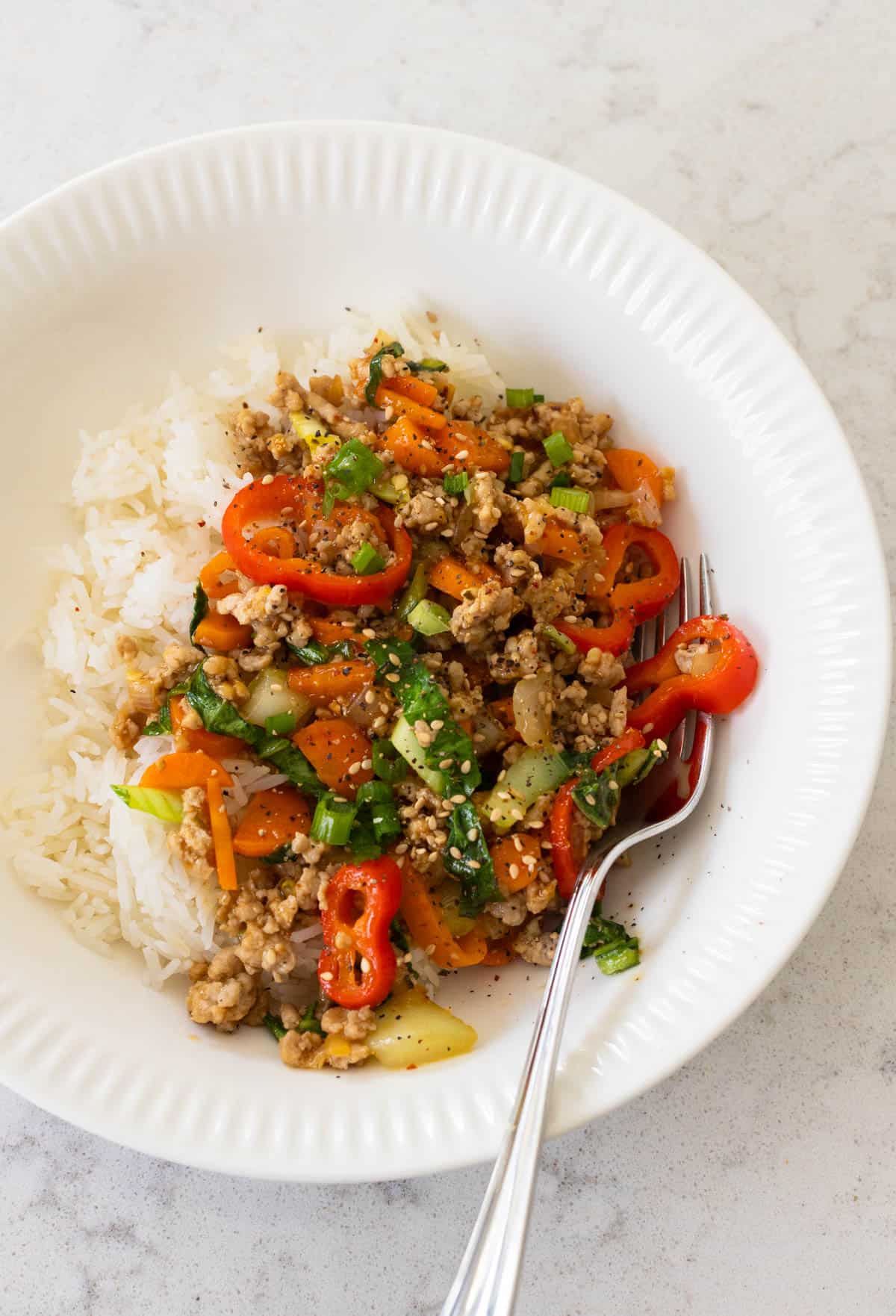 A white bowl has a bed of rice that's been topped with the ground pork stir fry loaded with peppers and cabbage.