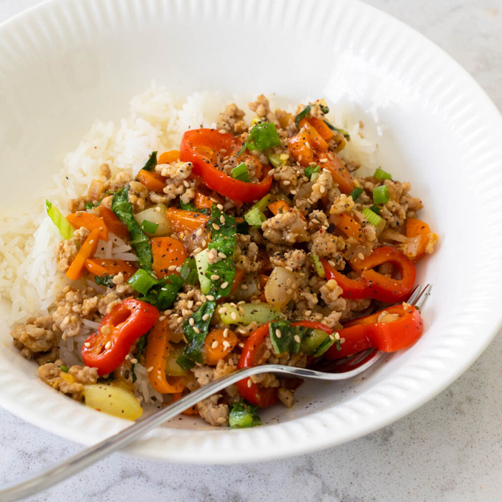A white bowl has a bed of rice that's been topped with the ground pork stir fry loaded with peppers and cabbage.
