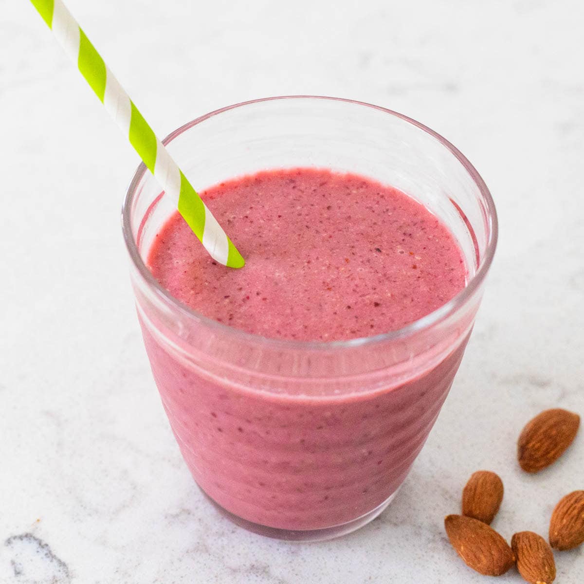A red cherry almond smoothie in a cup with a striped straw has almonds scattered on the counter next to it.