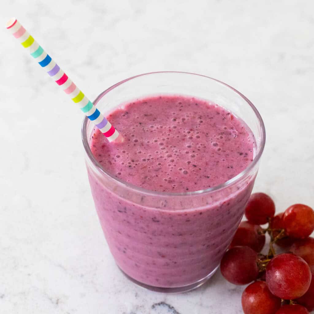 A deep purple grape smoothie in a cup with a straw and a cluster of grapes on the side.