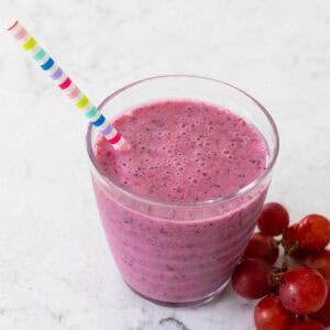 A deep purple grape smoothie in a cup with a straw and a cluster of grapes on the side.