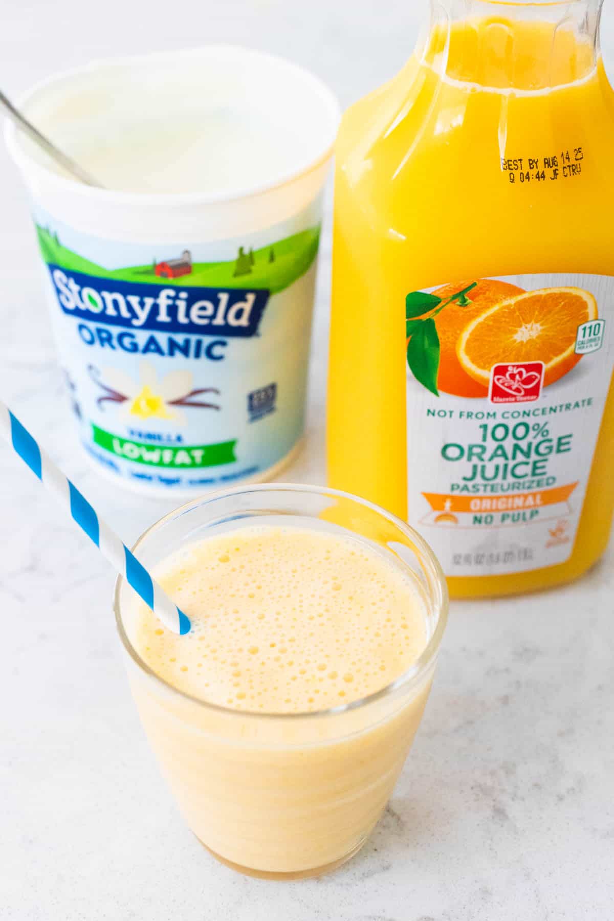 The smoothie is in a cup with a straw, the bottle of orange juice and container of yogurt sit behind it.