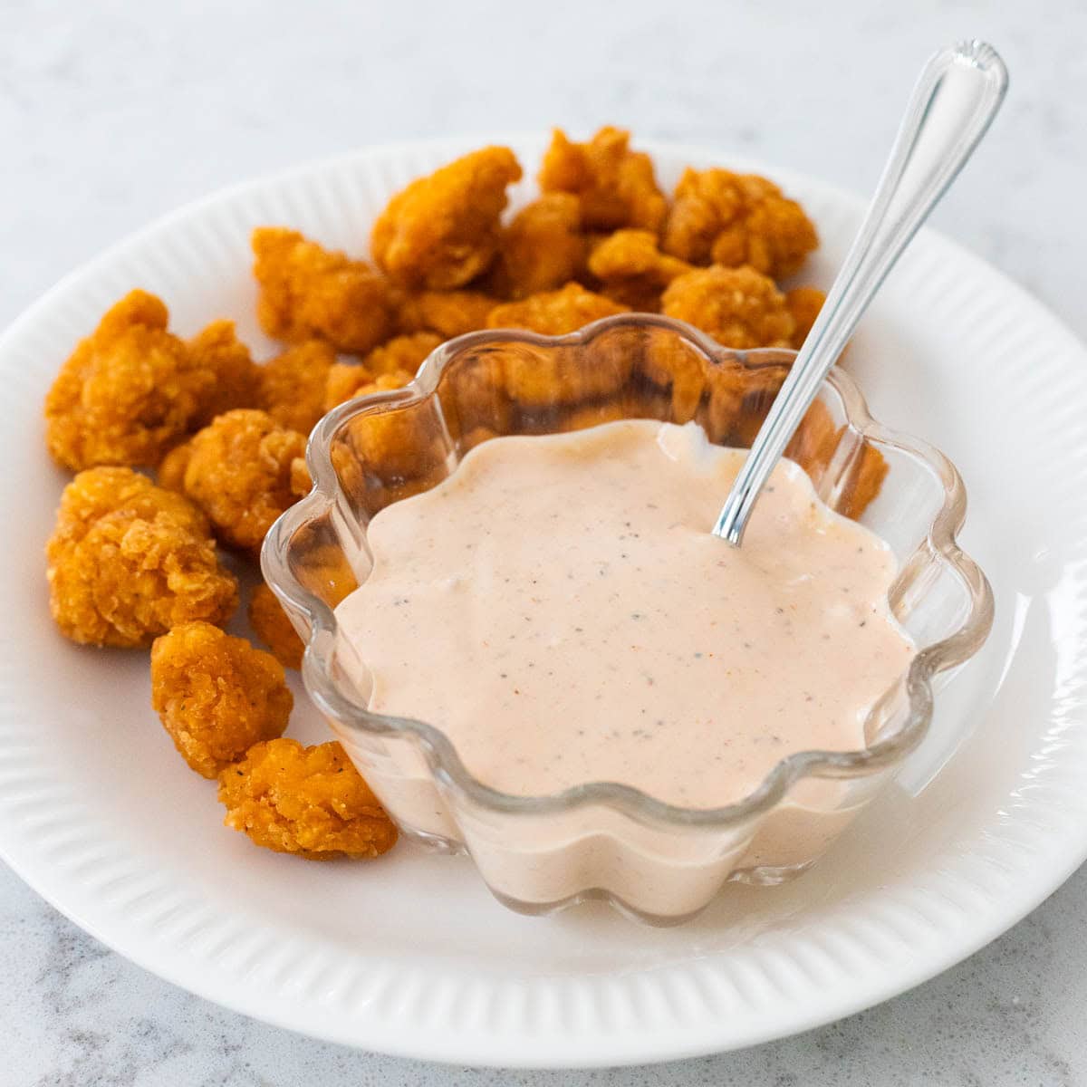 A bowl of sauce with a spoon sits on a plate of popcorn chicken.