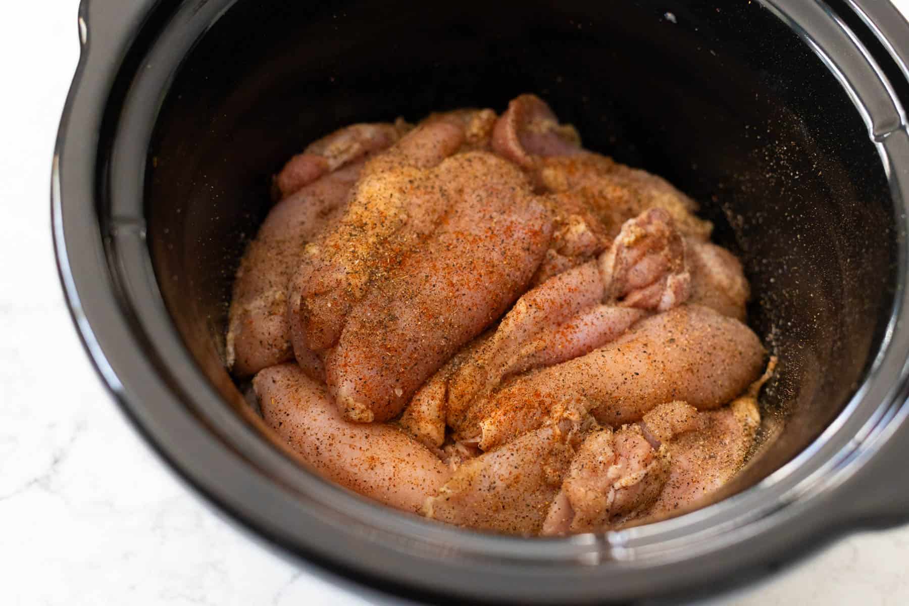 The raw chicken is sitting in the crockpot with spices on them.