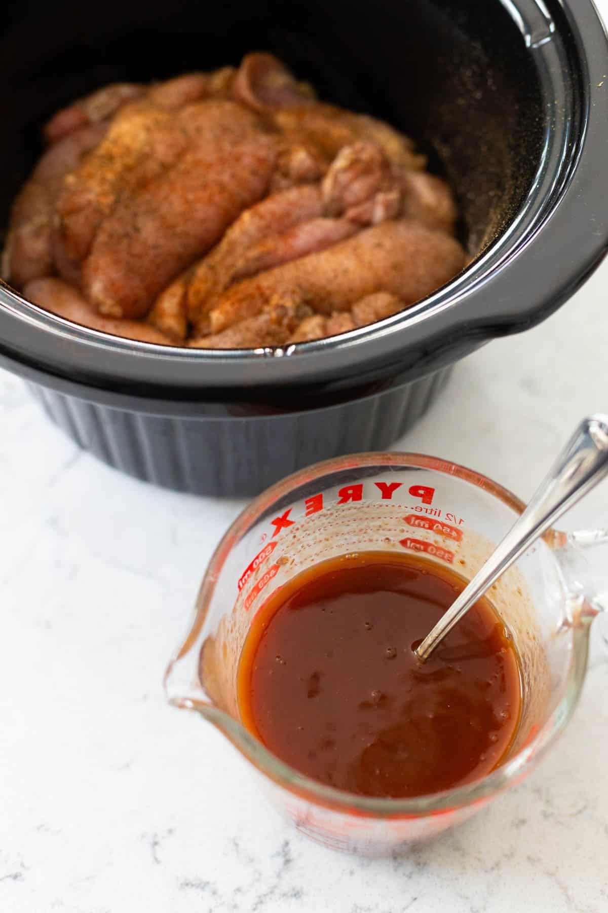A liquid measuring cup filled with the BBQ peach sauce is sitting next to the crockpot of raw chicken.