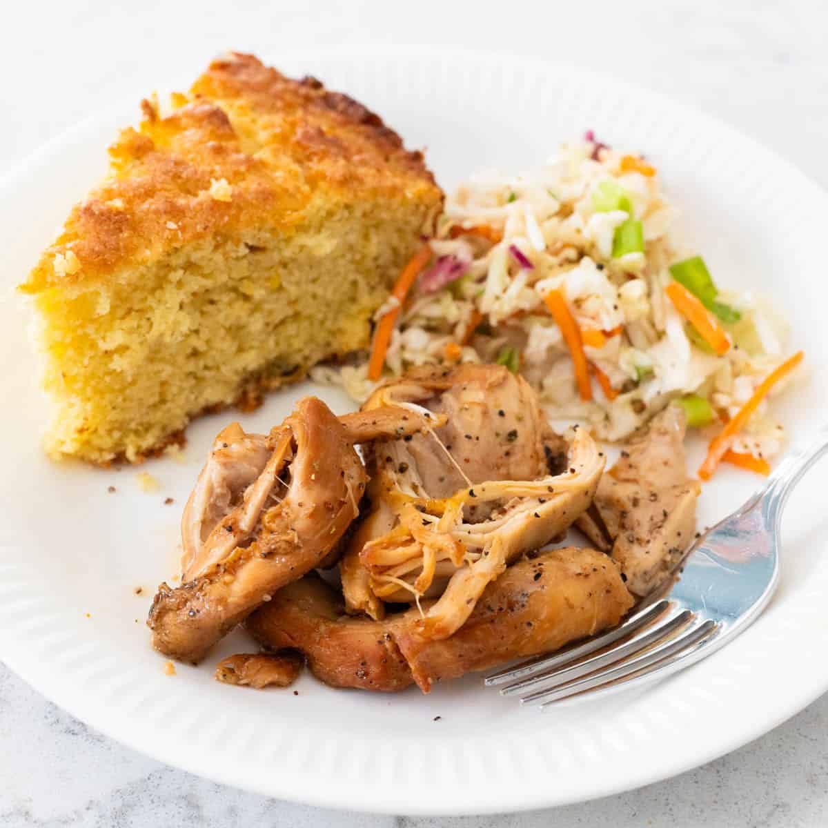 A scoop of the BBQ peach chicken is on a white plate with a slice of fresh cornbread and scoop of vinegar coleslaw.