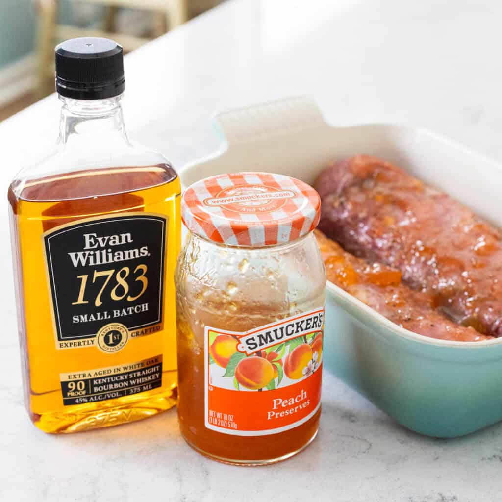 A bottle of bourbon and a jar of peach preserves sits next to a dish of pork.