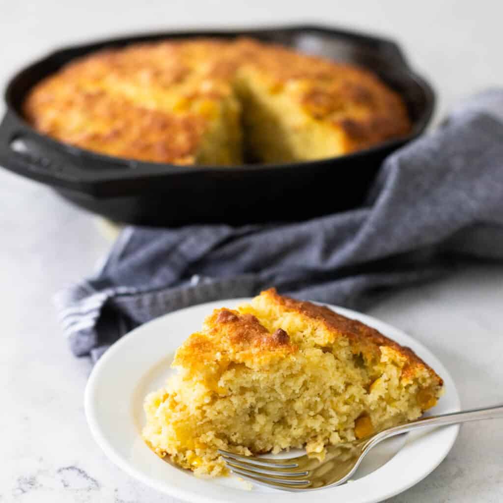 A wedge of homemade cornbread sits on a plate in front of the cast iron skillet it was baked in.