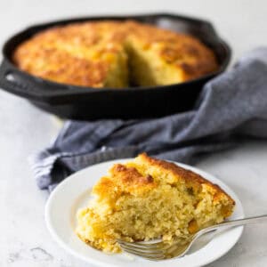 A wedge of homemade cornbread sits on a plate in front of the cast iron skillet it was baked in.