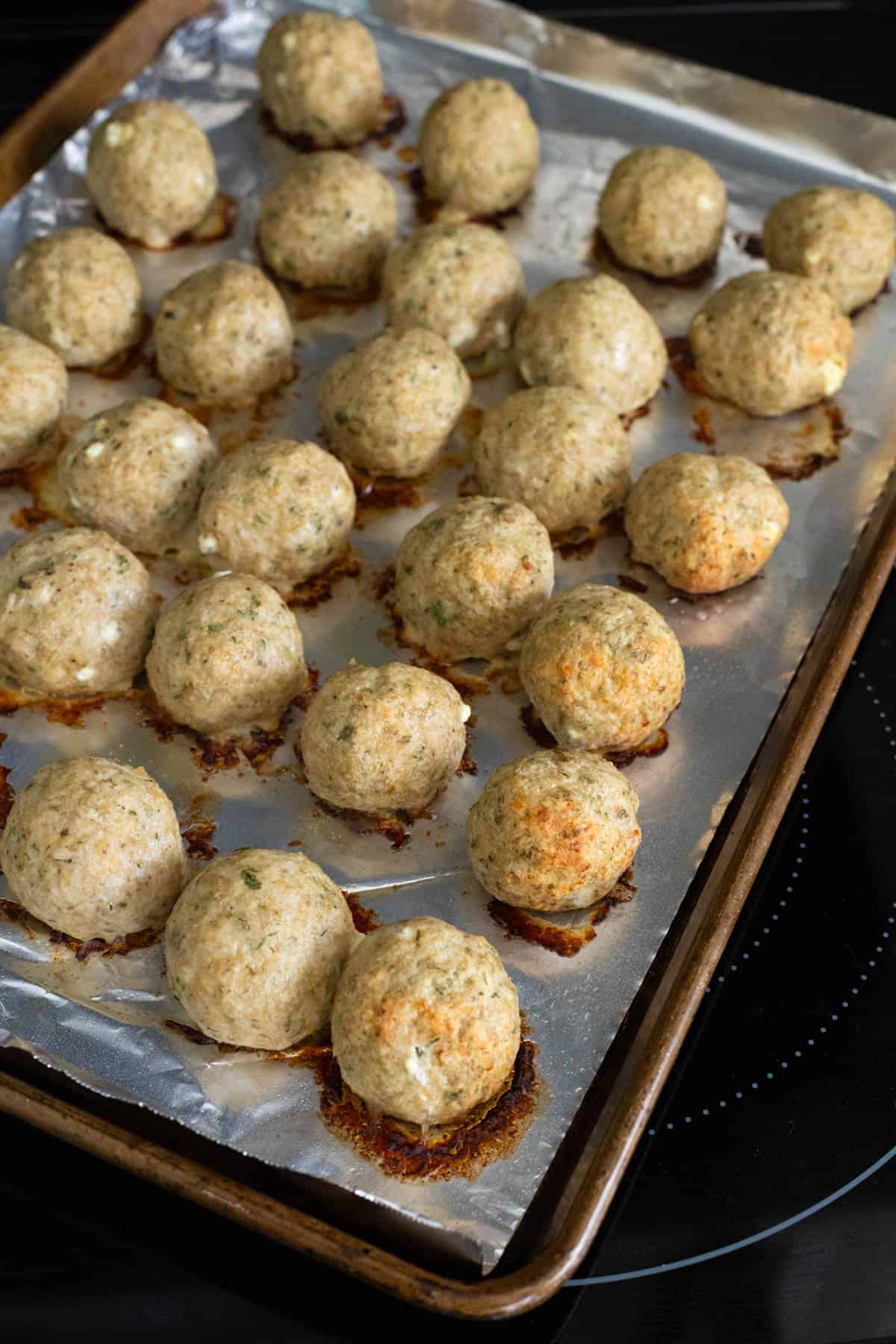 A baking sheet on the stovetop has rows of baked gyro meatballs on it.