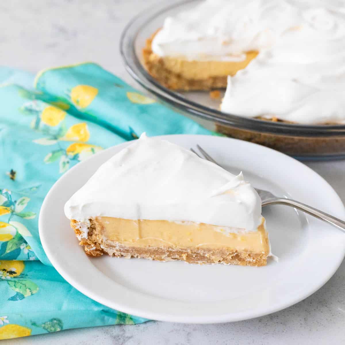 The slice of lemon pie with whipped topping is on a white plate next to a blue napkin with lemons on it.