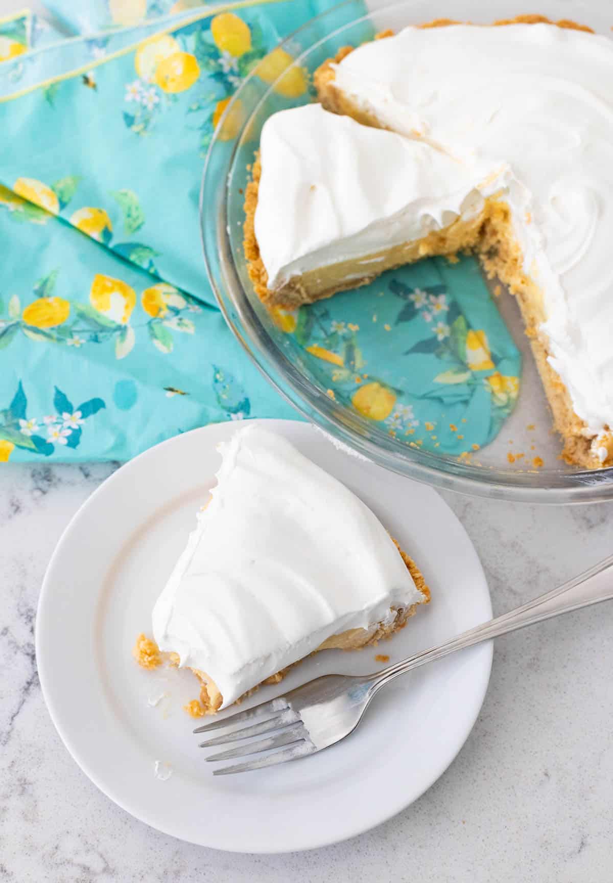The slice of lemon pie with whipped topping is on a white plate next to a blue napkin with lemons on it.