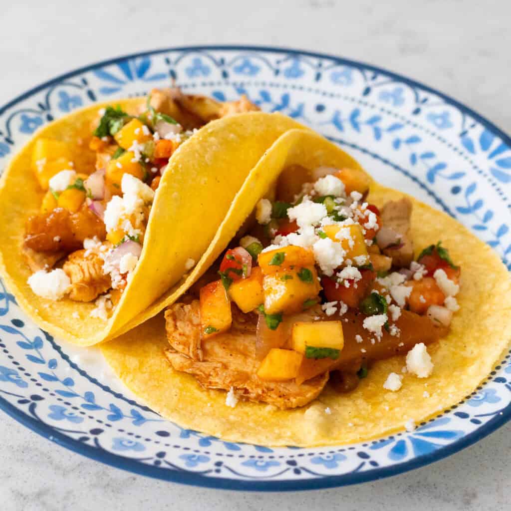 Two tacos topped with peach salsa and crumbled cheese are on a blue and white plate.