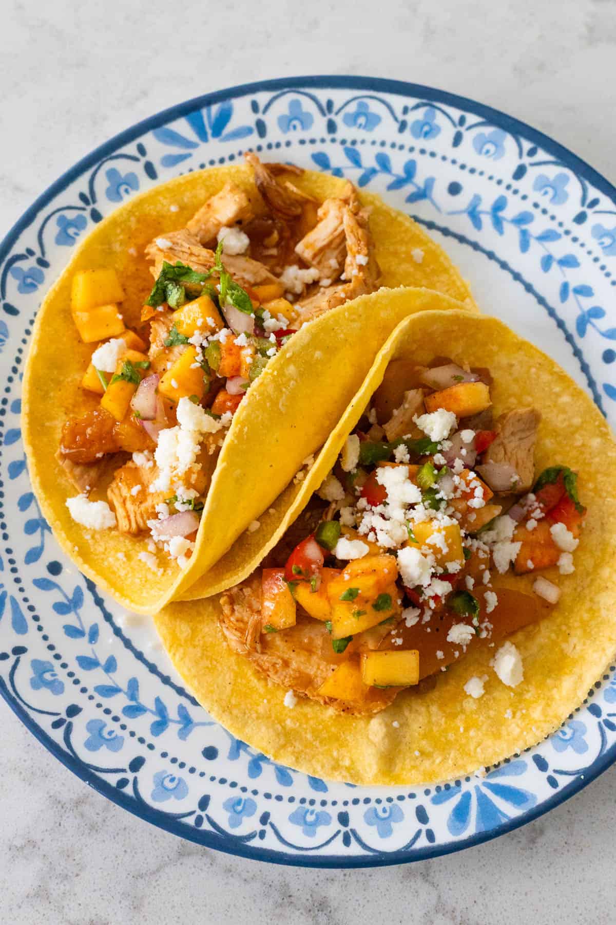 Two tacos topped with peach salsa and crumbled cheese are on a blue and white plate.