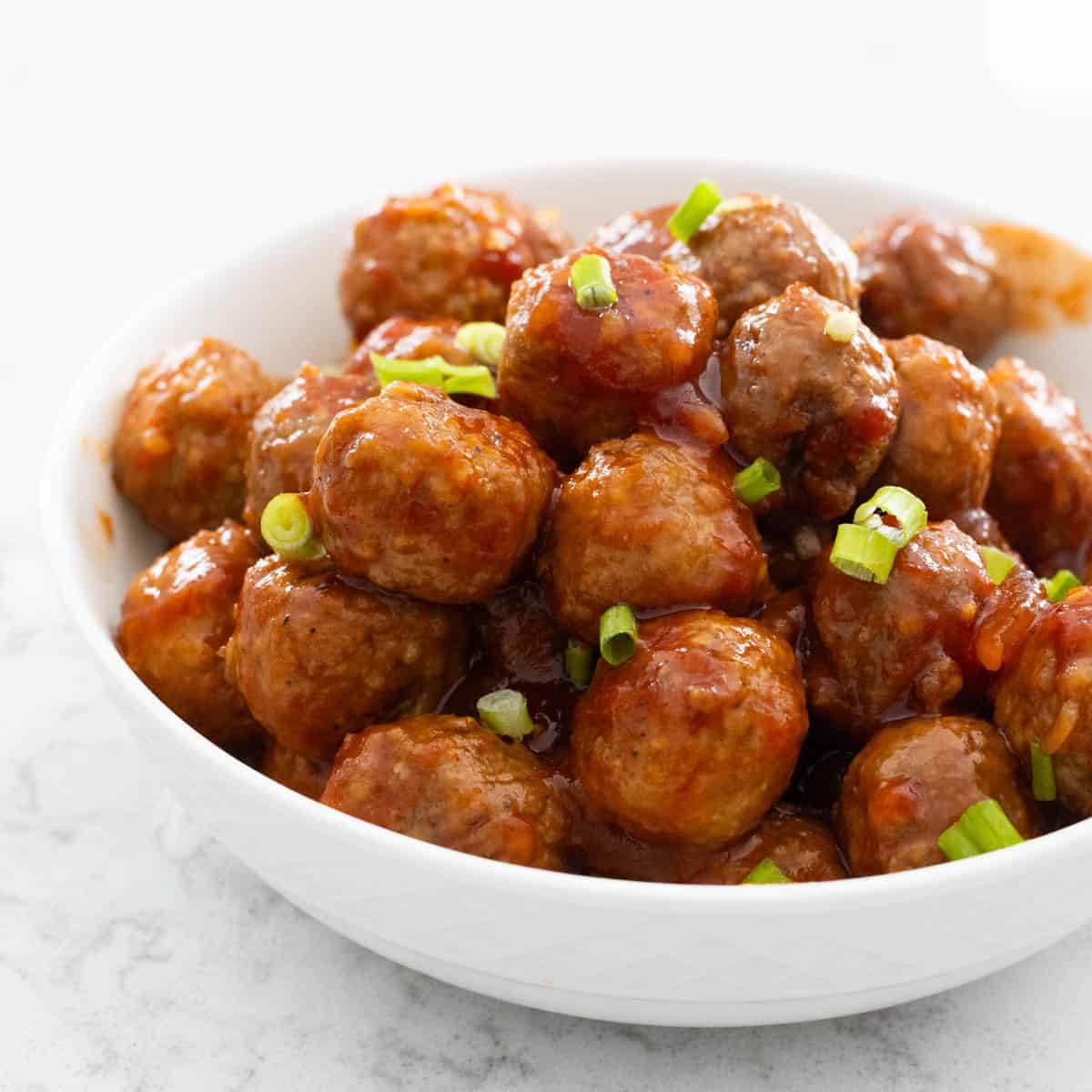 A white bowl is filled with glazed meatballs that have a sprinkle of chopped green onions on top for garnish.