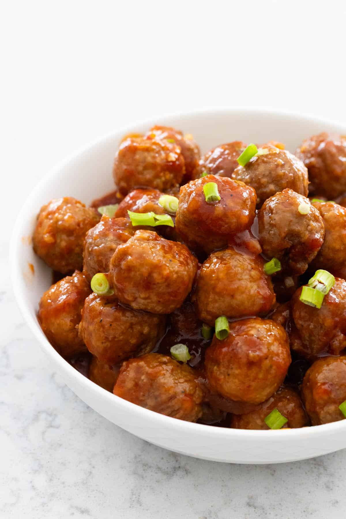 A white bowl is filled with glazed meatballs that have a sprinkle of chopped green onions on top for garnish.