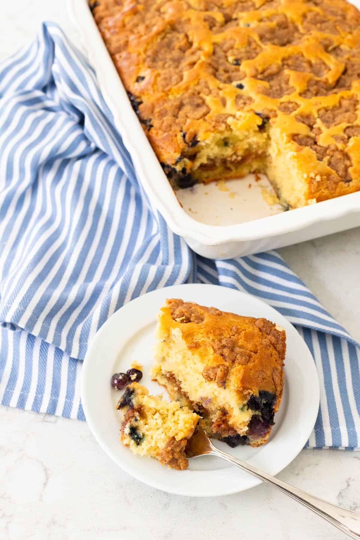 A square of fresh baked coffee cake with a layer of blueberries sits on a plate next to a full pan of the cake.