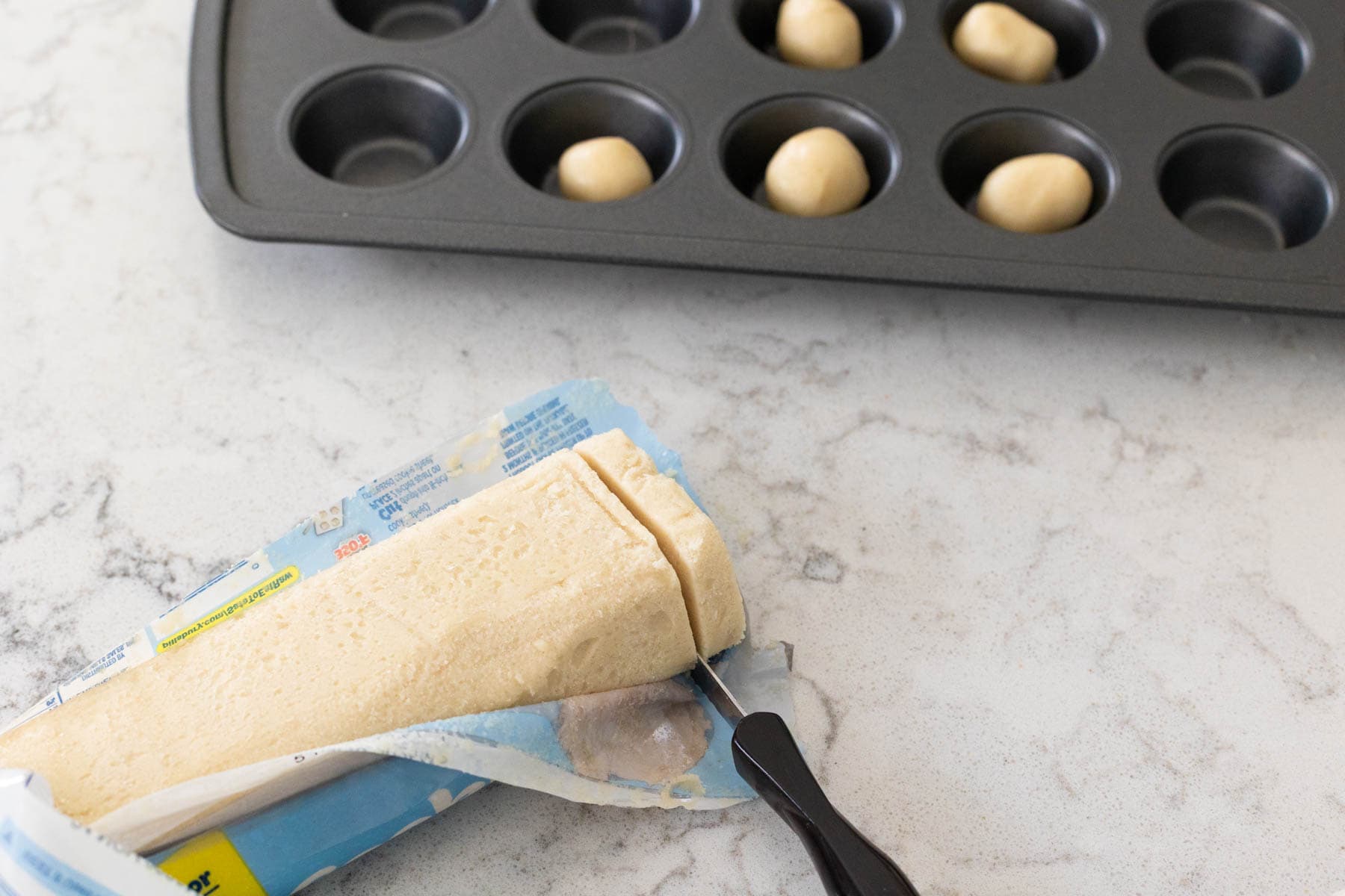 A block of refrigerated sugar cookie dough being sliced into pieces, with small dough balls placed in a mini muffin tin in the background.