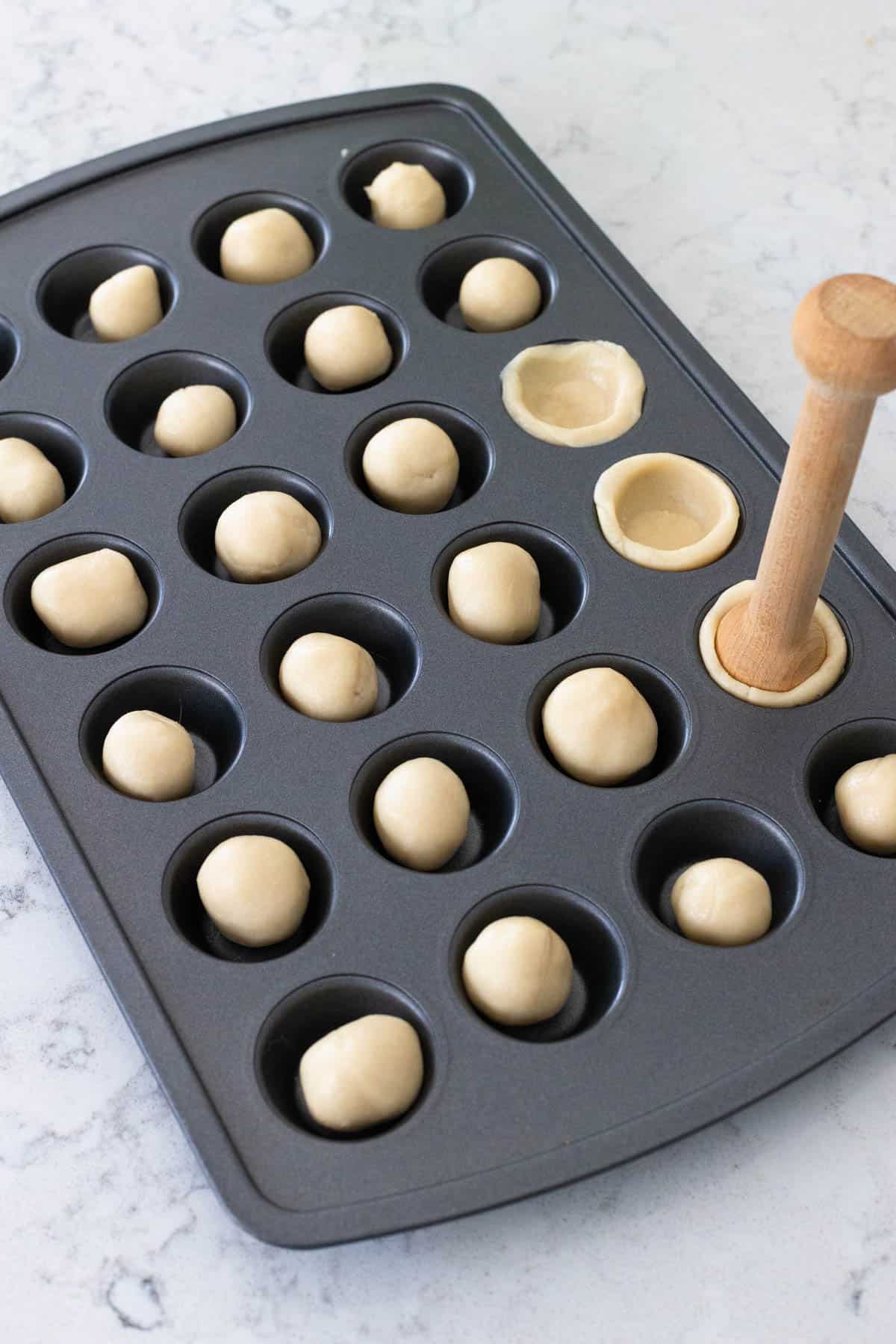 Sugar cookie dough balls placed in a mini muffin tin, with a wooden tool pressing some of them into cup shapes.