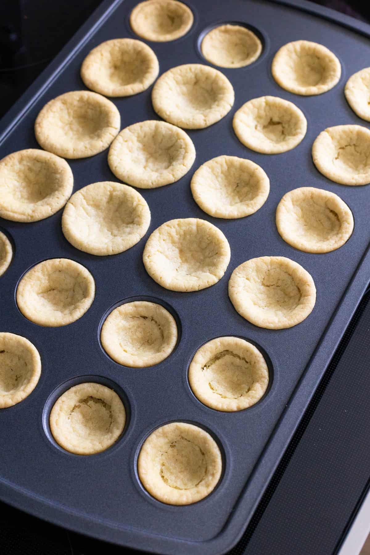A mini muffin tin filled with freshly baked sugar cookie cups, lightly golden with hollow centers ready for filling.