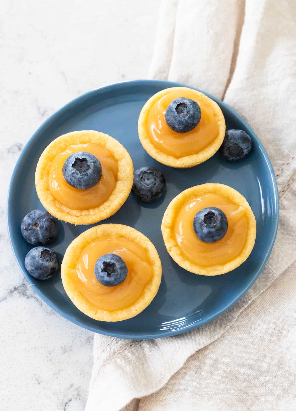A blue plate has little lemon curd filled sugar cookie cups with fresh blueberries on top.