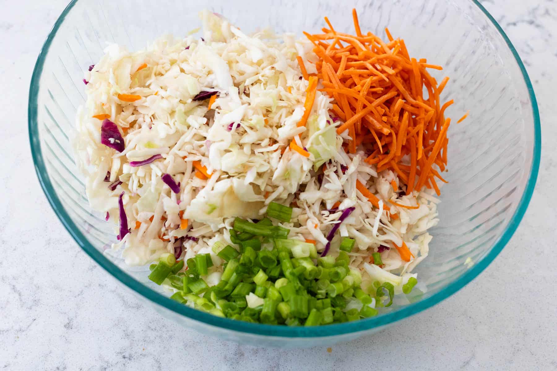 A large mixing bowl is filled with the different vegetables needed for the slaw that are ready to be mixed together.