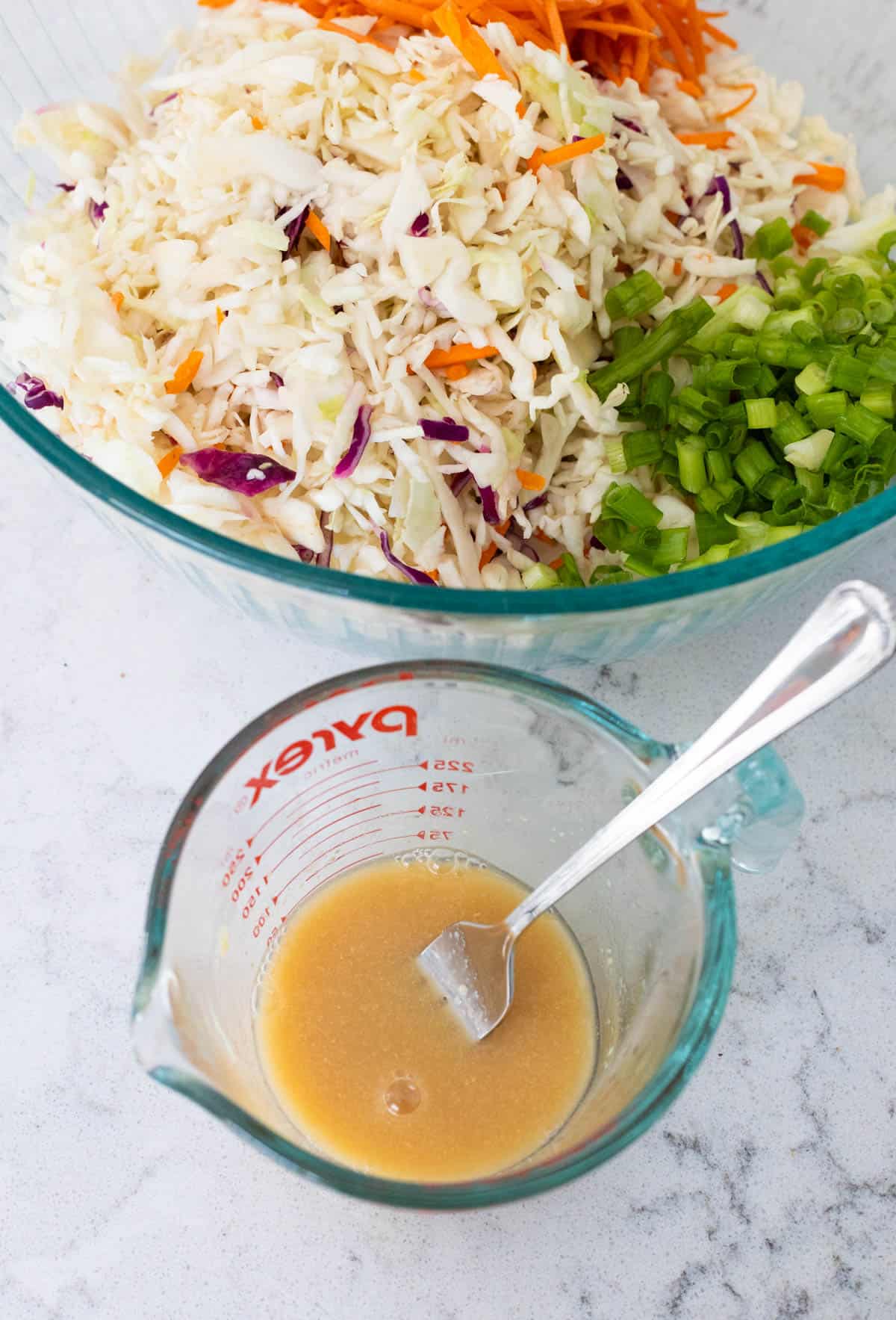 The vinegar dressing is in a liquid measuring cup next to the large mixing bowl filled with slaw.