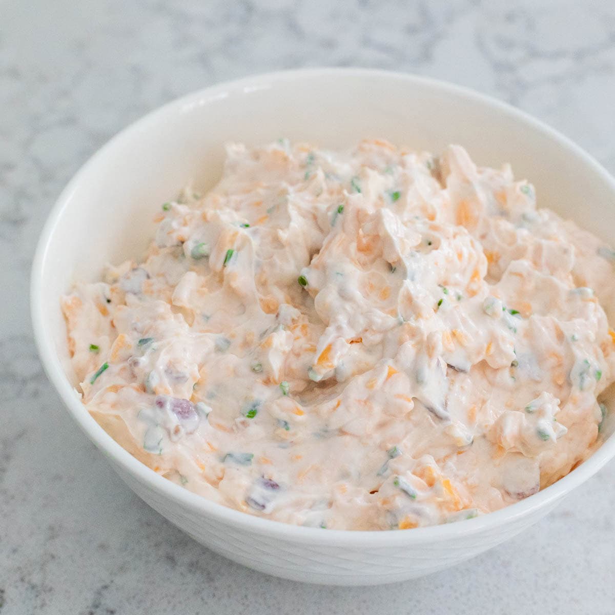 A creamy mixture of shredded cheddar cheese, sour cream, crumbled bacon, and chives in a white bowl, ready to serve as a dip.