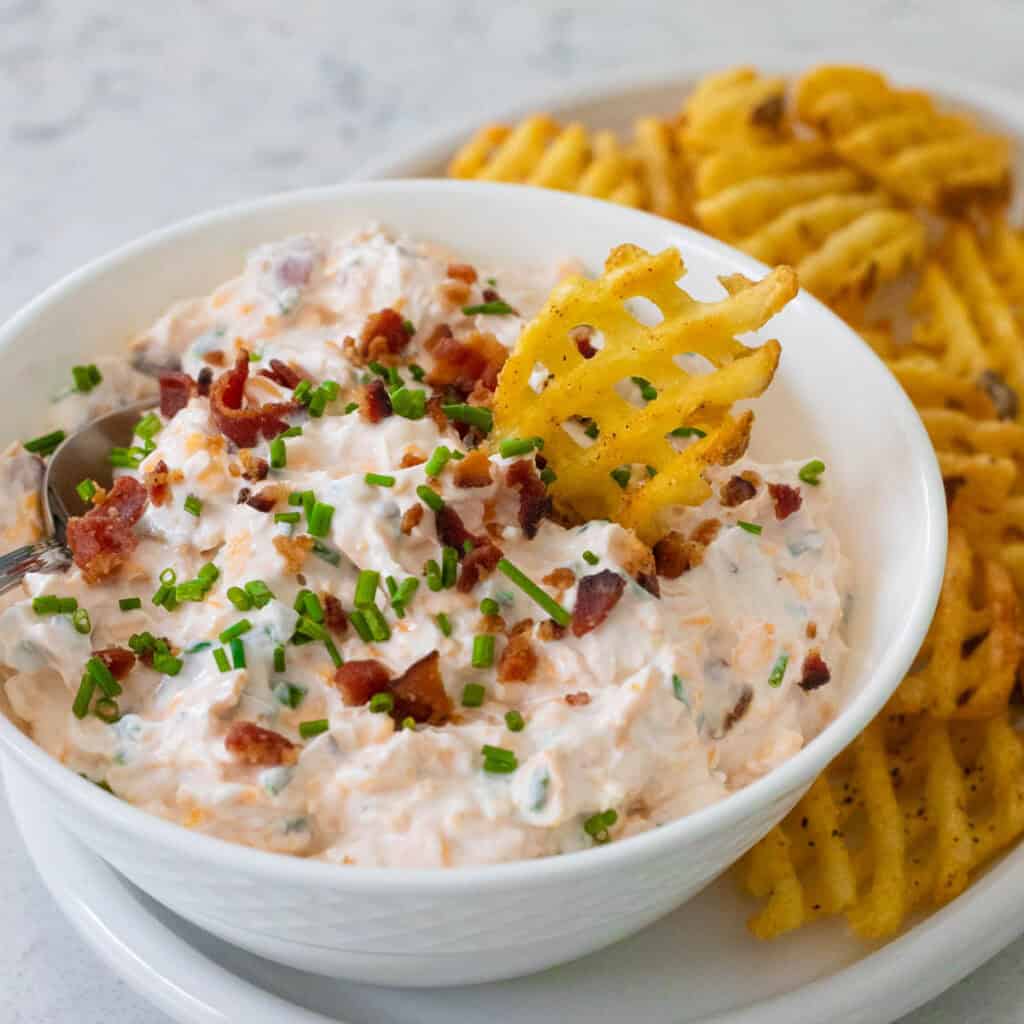 A bowl filled with a sour cream, bacon, and cheese dip is topped with chopped fresh chives. Baked waffles fries are on the platter next to the bowl.