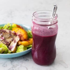 A mason jar filled with deep purple blueberry vinaigrette sits on the counter next to a fresh peach salad.