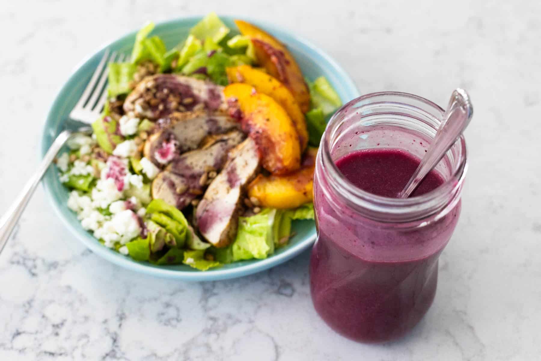 A mason jar filled with deep purple blueberry vinaigrette sits on the counter next to a fresh peach salad.