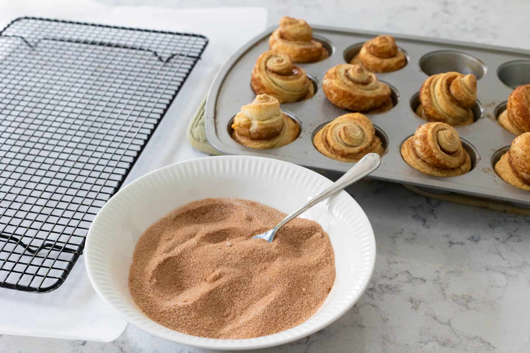 Freshly baked cruffins in a muffin tin next to a bowl of cinnamon sugar and a cooling rack, ready for coating.