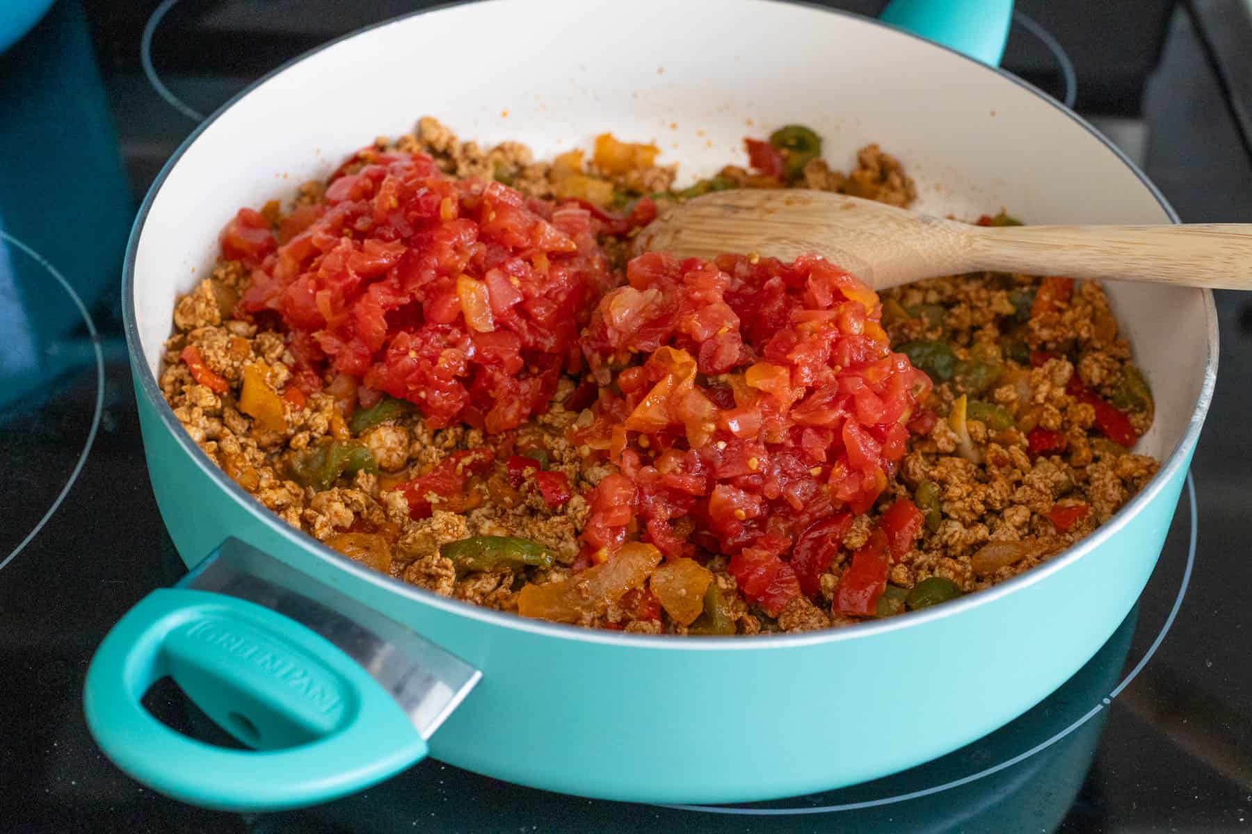 Ground meat, veggies, and diced tomatoes in a turquoise skillet on the stove, with a wooden spoon.