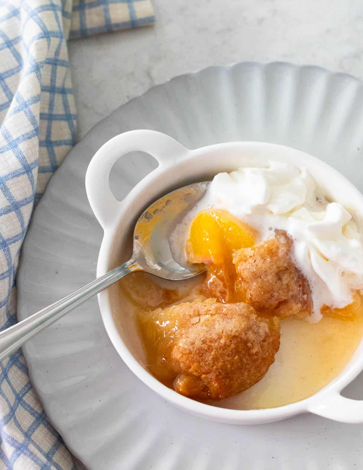 A single peach dumpling is topped with whipped cream and has a spoon on the side.