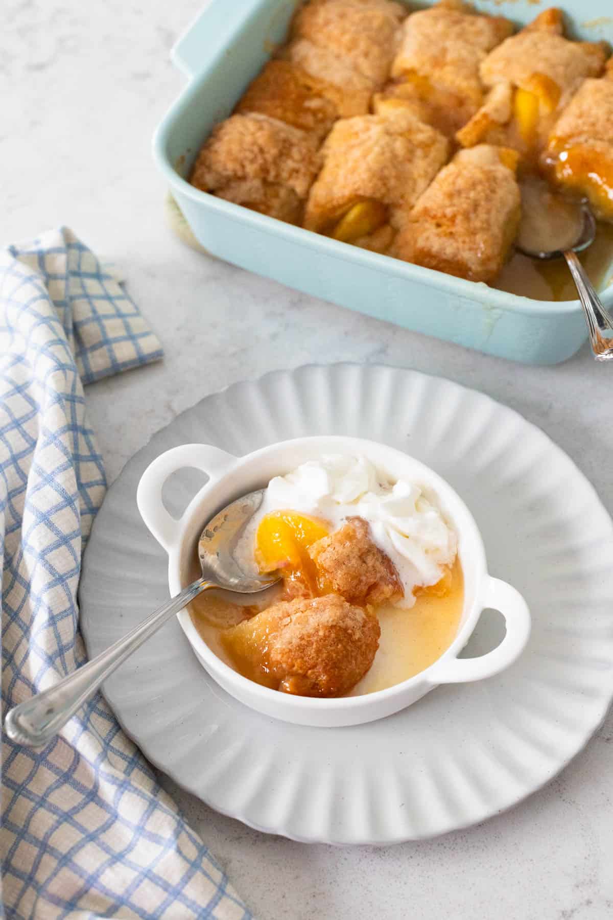 A warm peach dumpling topped with whipped cream sits on a ruffled plate. The baking dish filled with dessert is in the back.