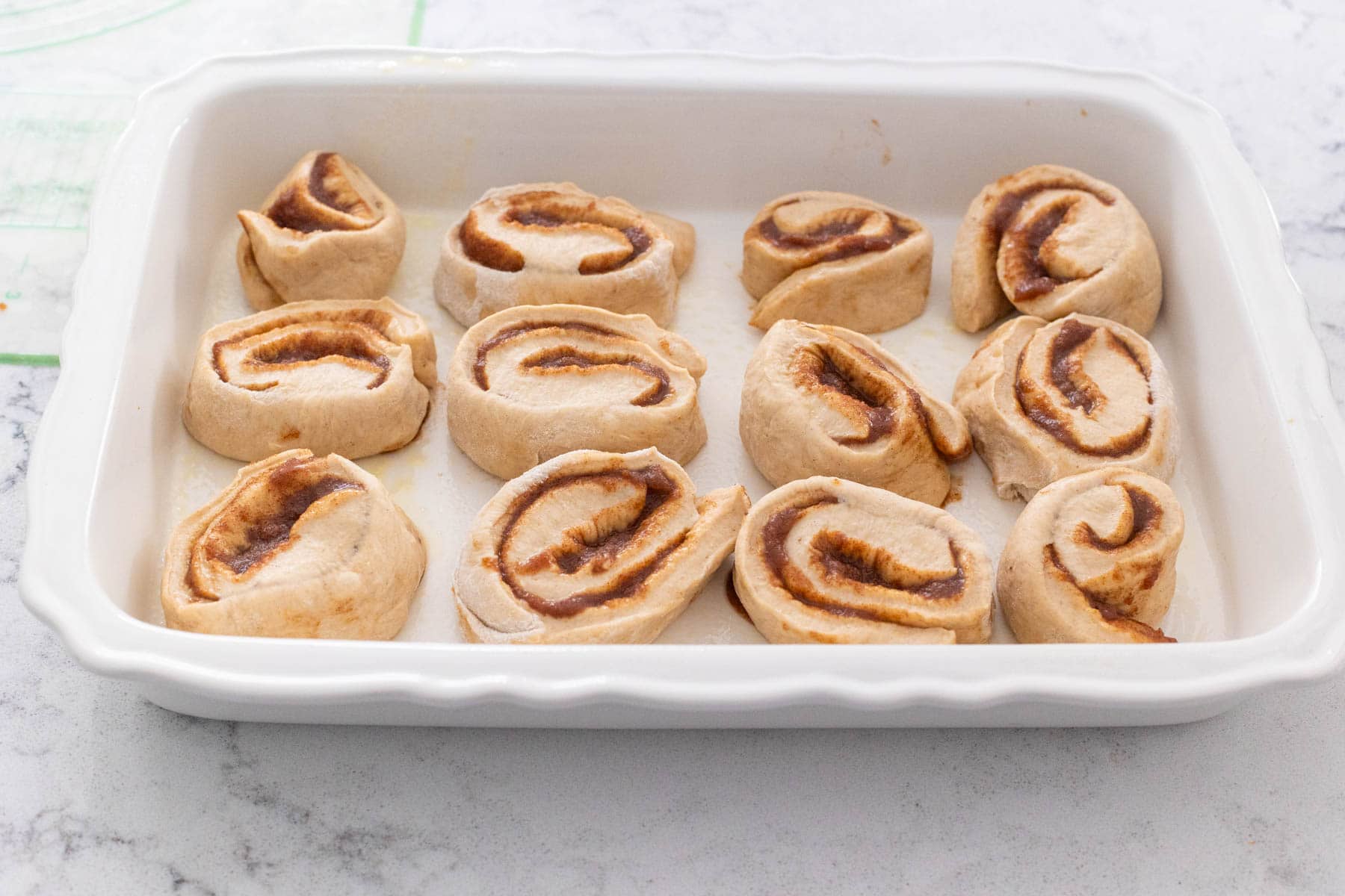 Unbaked apple cider cinnamon rolls arranged in a white ceramic baking dish, showing swirls of cinnamon filling before the final rise.