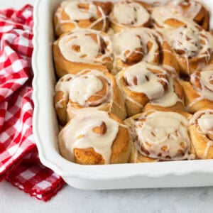 A white baking dish of apple cinnamon rolls with maple glaze sits next to a red and white napkin.