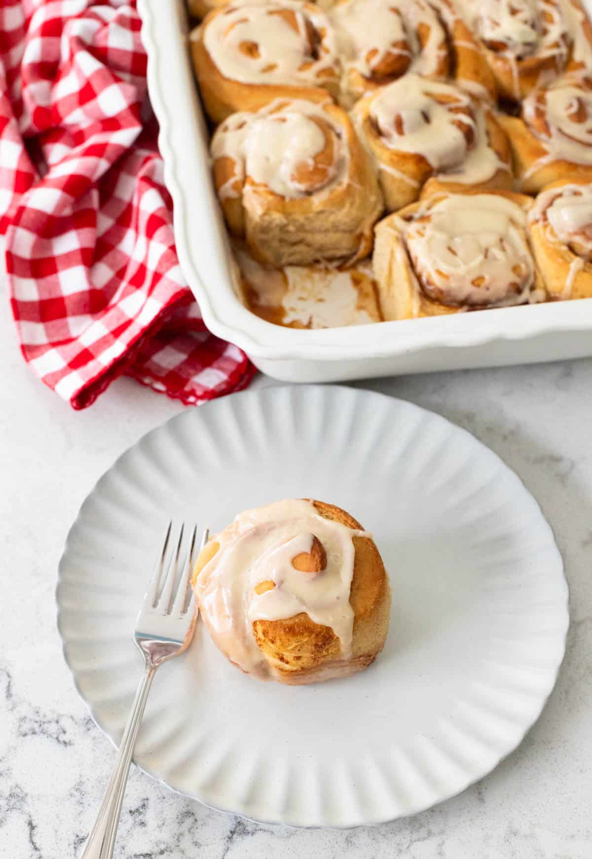 A dish of freshly baked apple cider cinnamon rolls topped with glaze, with one roll served on a white plate alongside a fork.