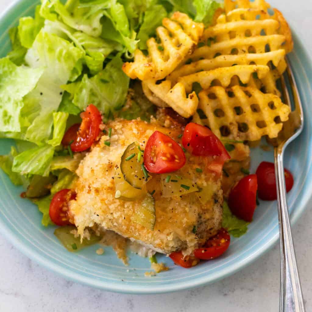 A breaded chicken baked with Chick-Fil-A sauce is topped with chopped pickles and tomatoes and served with a side salad and baked waffles fries.