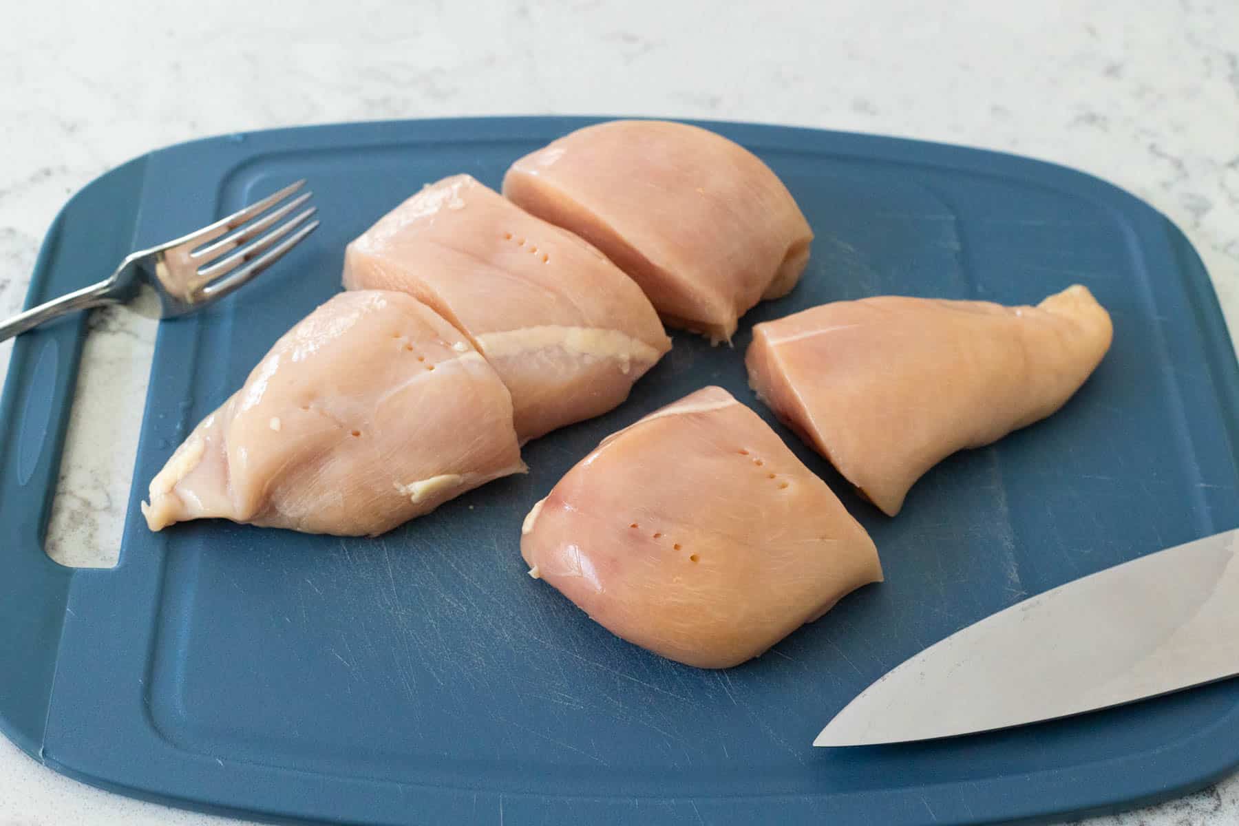 Raw chicken breasts cut into pieces on a blue cutting board with a knife and fork.