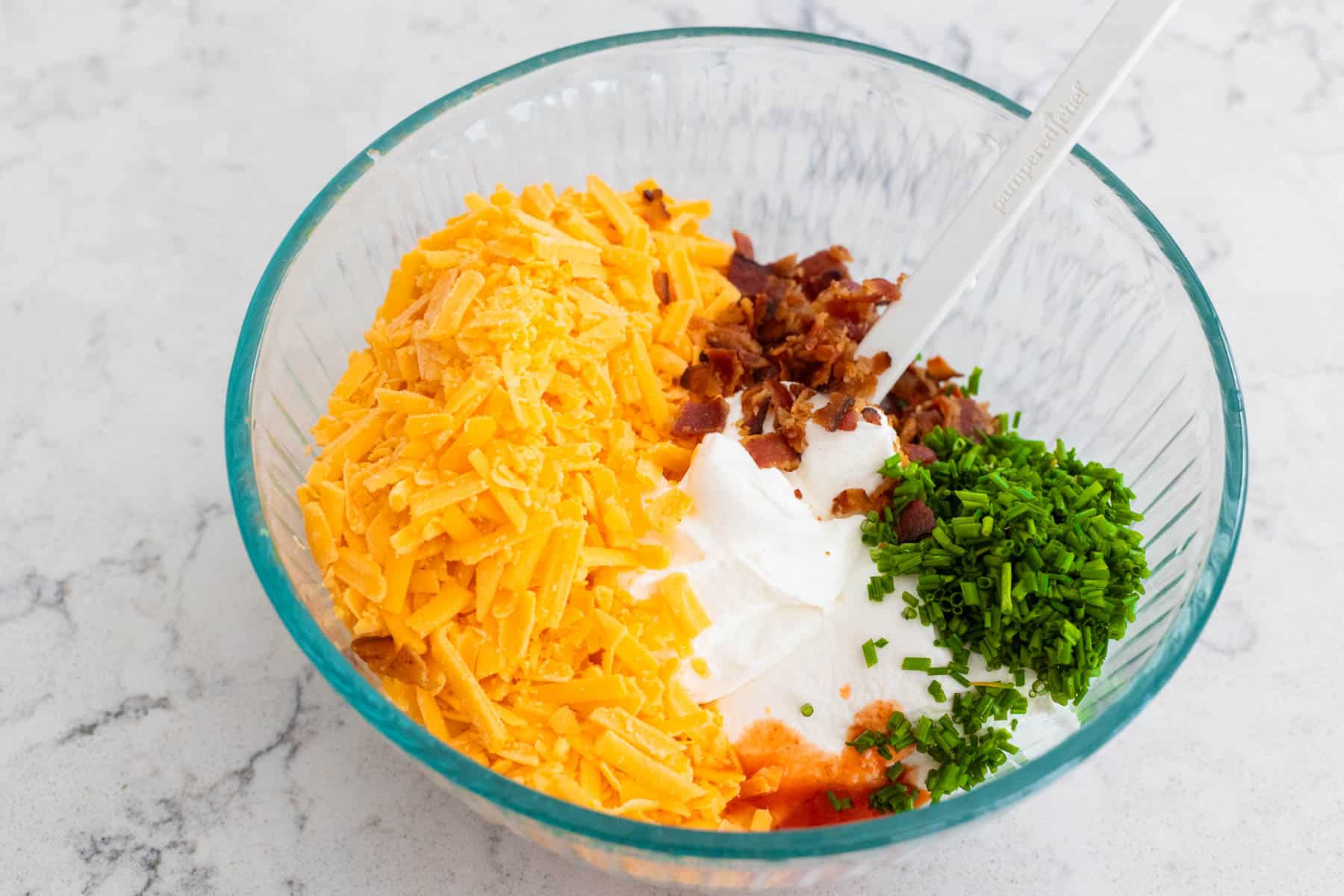 A glass mixing bowl containing shredded cheddar cheese, sour cream, crumbled bacon, and chopped chives, with a spoon mixing the ingredients.