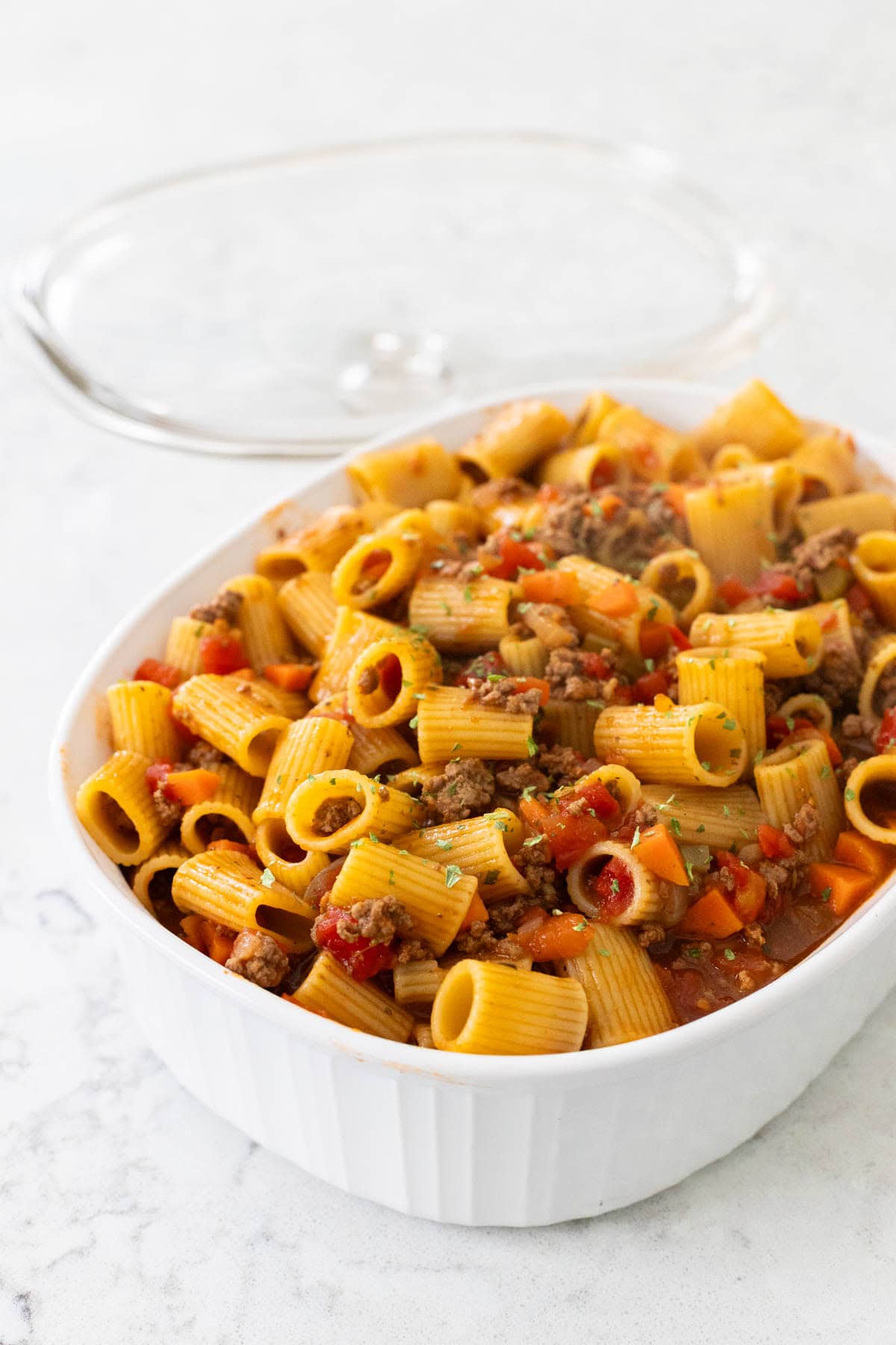 A serving of beef rigatoni pasta with diced tomatoes, and carrots, garnished with parsley in a white baking dish.