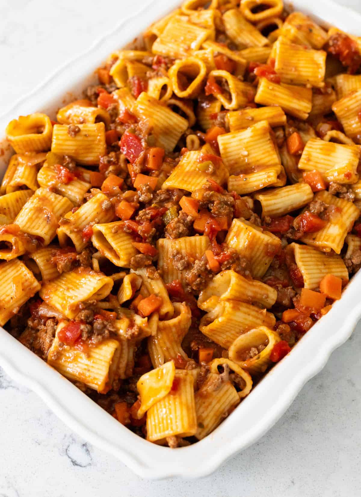 A serving of beef rigatoni pasta with diced tomatoes, and carrots, garnished with parsley in a white baking dish.