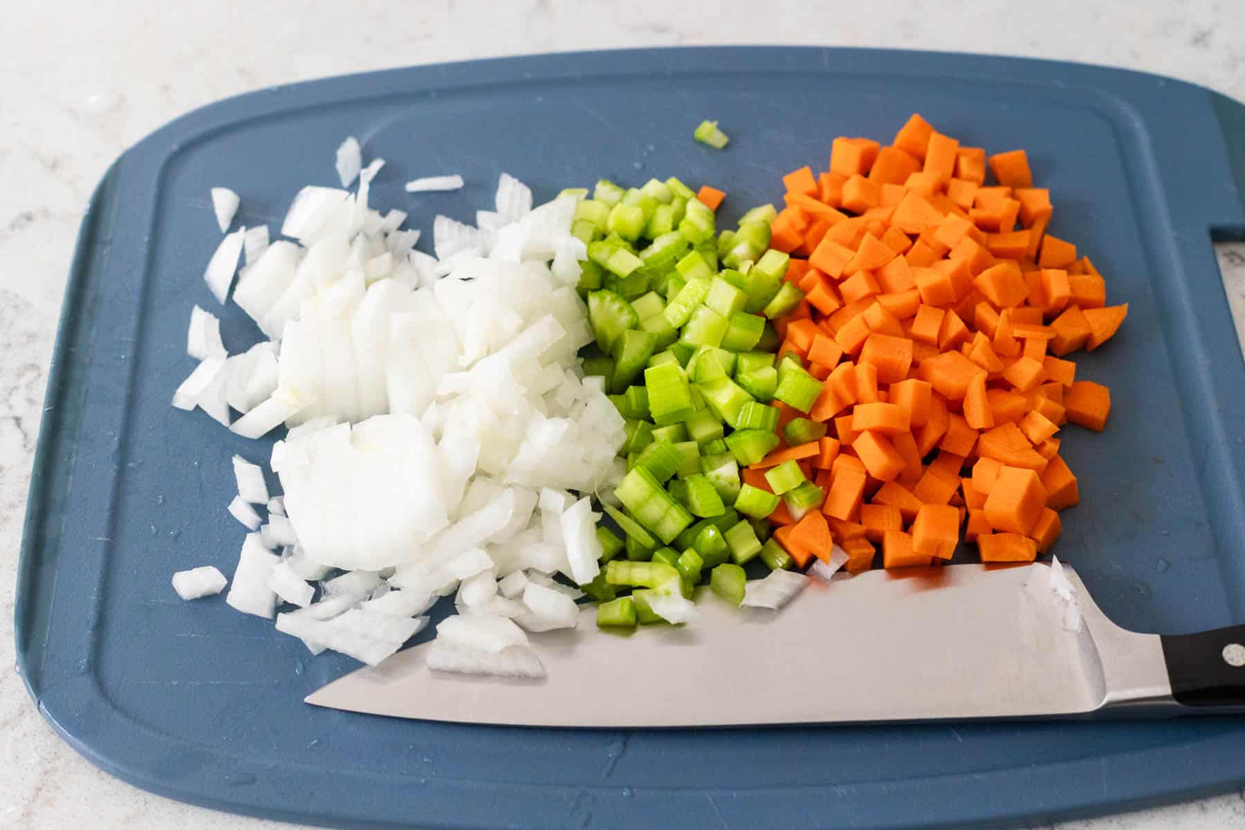 Chopped onions, celery, and carrots on a cutting board with a knife.